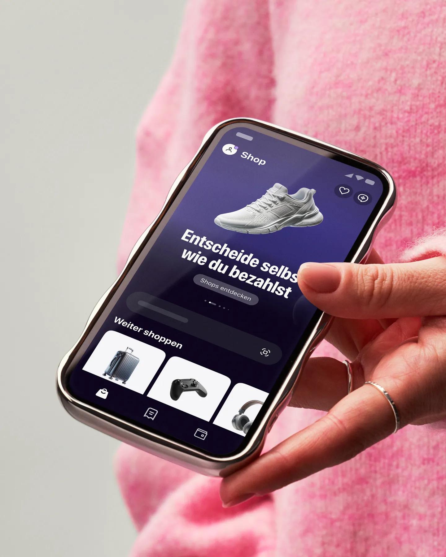 Hand holding a phone showing the Klarna app with pink sweater on background