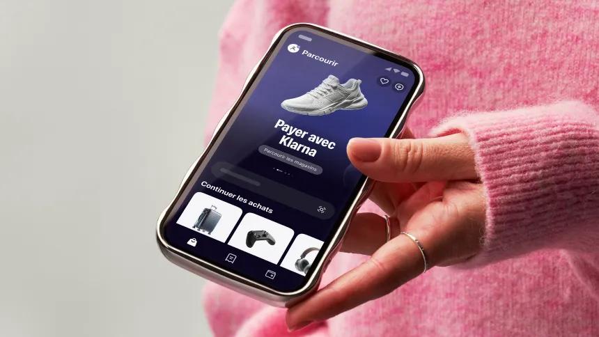 Hand holding a phone showing the Klarna app with pink sweater on background