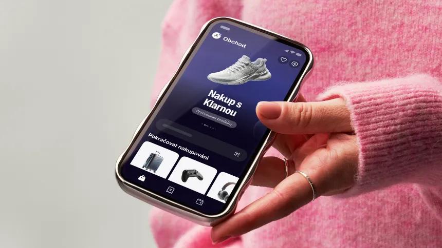 Hand holding a phone showing the Klarna app with pink sweater on background