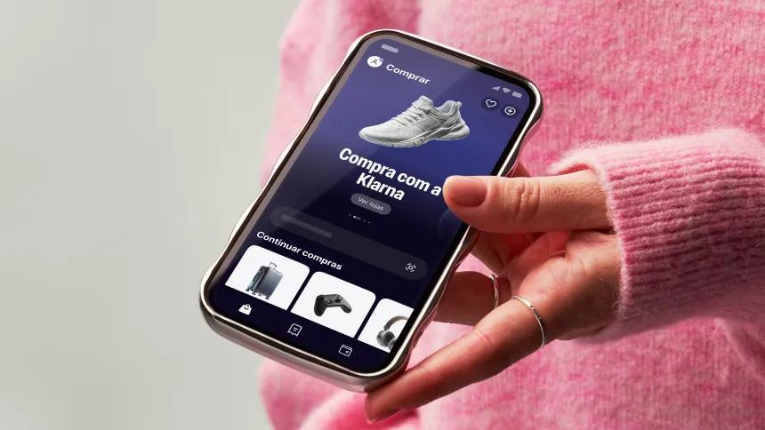 Hand holding a phone showing the Klarna app with pink sweater on background